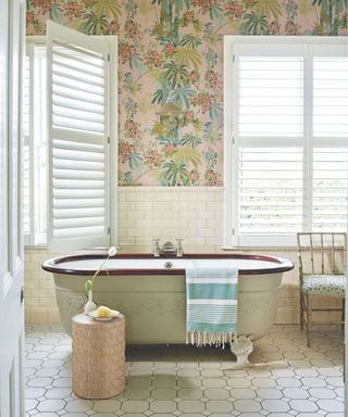 A colorful bathroom with pink leafy patterned wallpaper, white tiles, an ornate stand alone roll-top bath with a brown rim and blue and white striped towel, white tile floors, a small textured pink table with a single tulip in a white vase and a bamboo upholstered chair