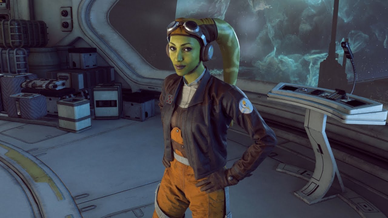 Hera Syndulla is a green-skinned Twi'lek with two head tails (known as lekku) protruding from the top left and right of her head. She is wearing a brown leather cap, goggles, a brown leather aviator jacket with a fluffy collar, and an orange jumpsuit. She is casually standing with her gloved hands on her hips inside a spaceship.