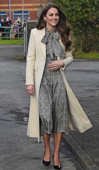 Kate Middleton wearing a black and white plaid dress and long white coat walking along a sidewalk