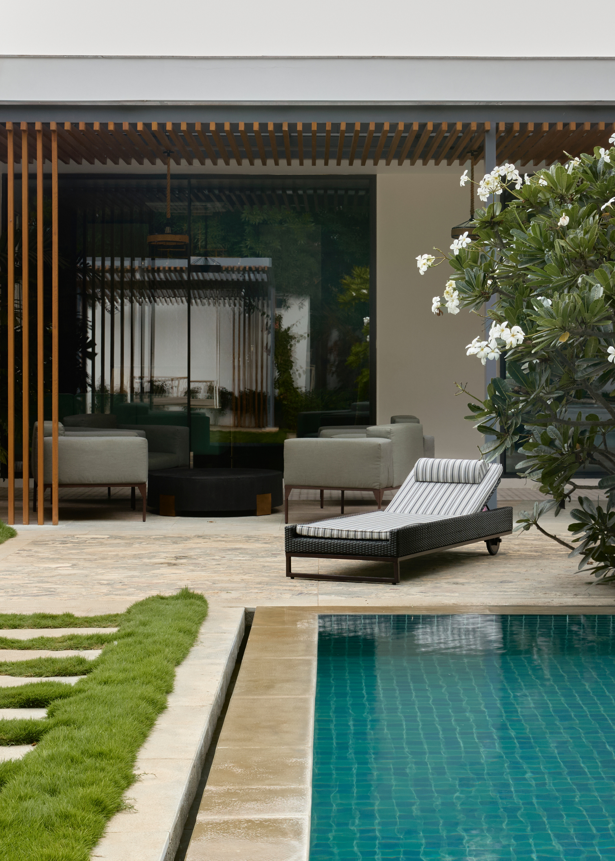 A garden with a frangipani tree, a blue pool, a striped sun bed and an outdoor living room