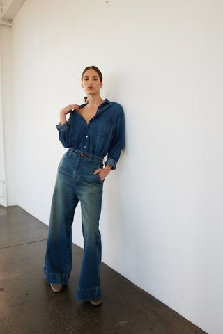 a model stands in front of a blank wall wearing a denim shirt from jamie haller with matching jeans and jamie haller shoes