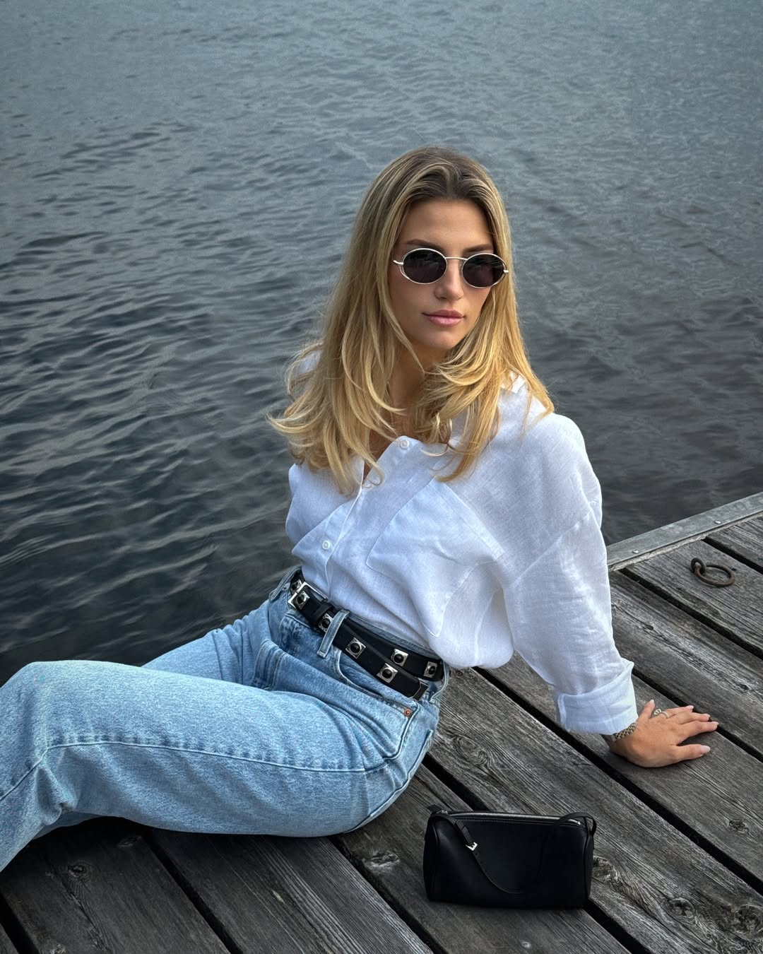 Cass Dimicco poses by the water with glossy strands, a white button-up, and blue jeans