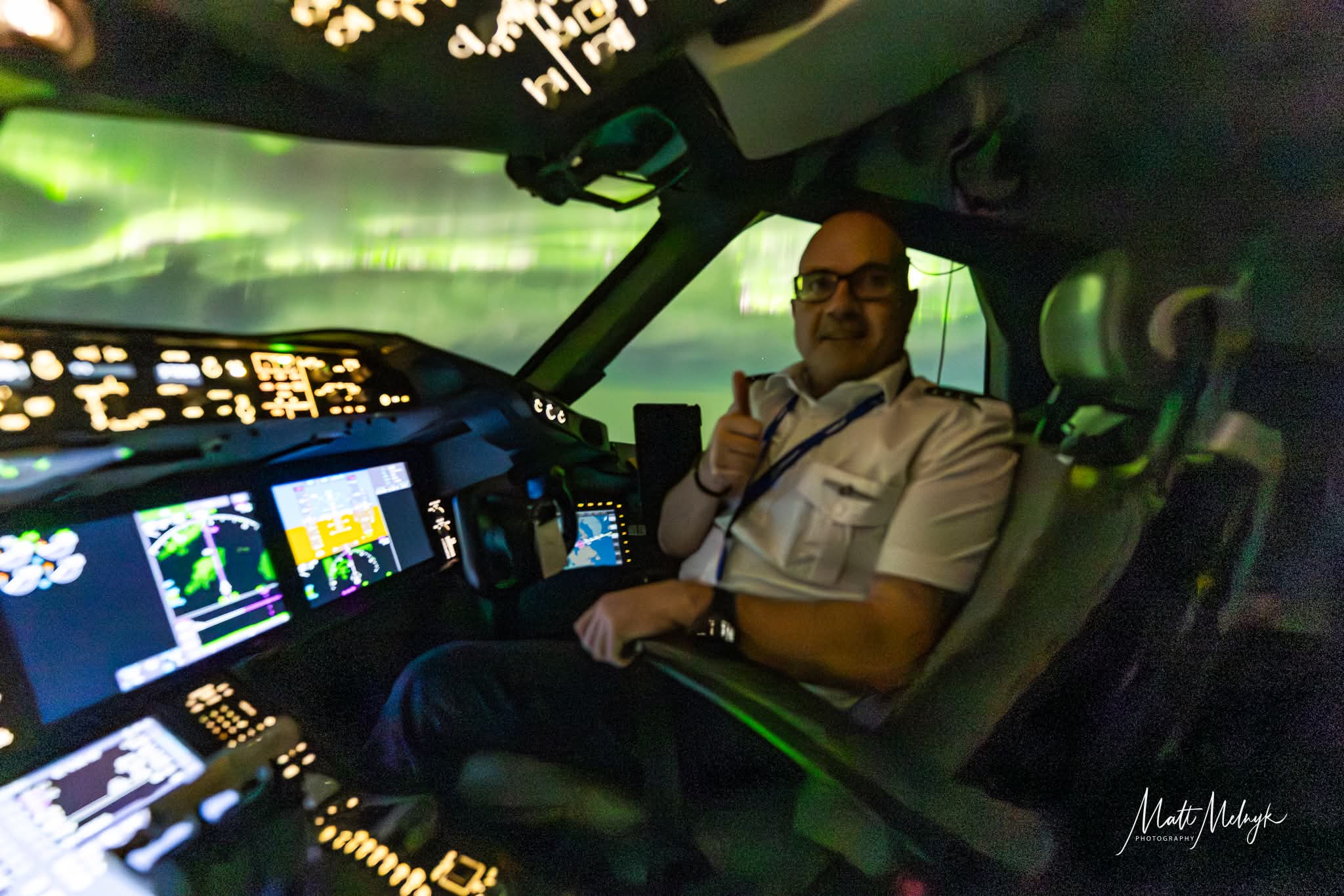 a man sits in the cockpit of a plane smiling and giving a thumbs up. out the window there are ribbons of green aurora light completely filling the sky.