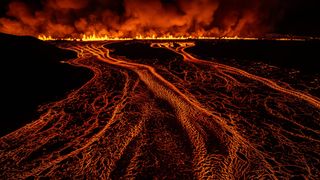Lava flows create bright, fiery patterns against a dark landscape, illuminated by flames and smoke rising into the night sky