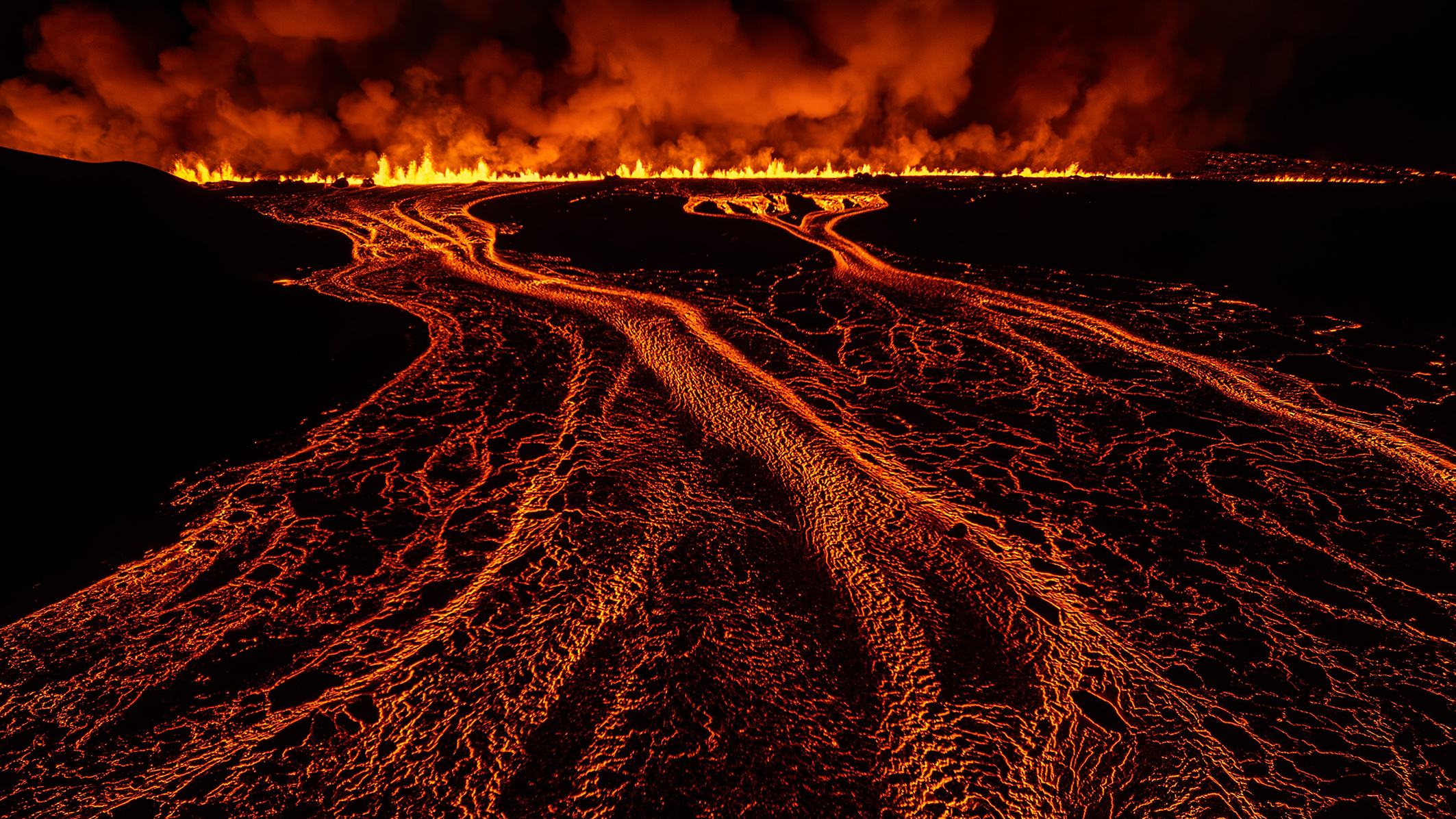 Lava flows create bright, fiery patterns against a dark landscape, illuminated by flames and smoke rising into the night sky