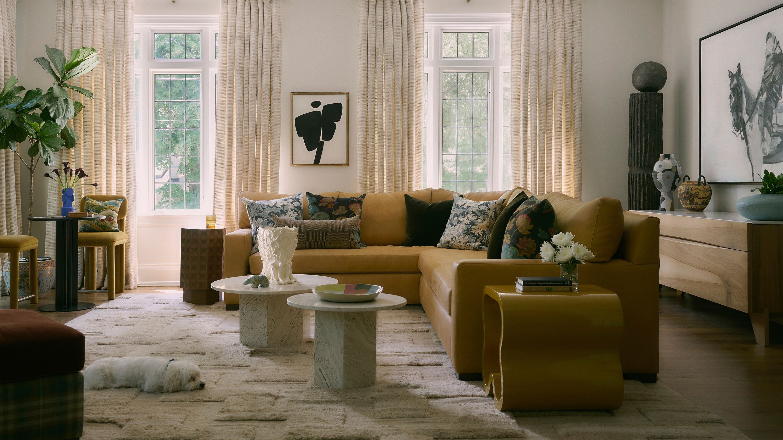 a large living room with neutral walls, large mustard yellow sectional at the center, eclectic art and objects, a neutral textured rug with a small dog lying on it