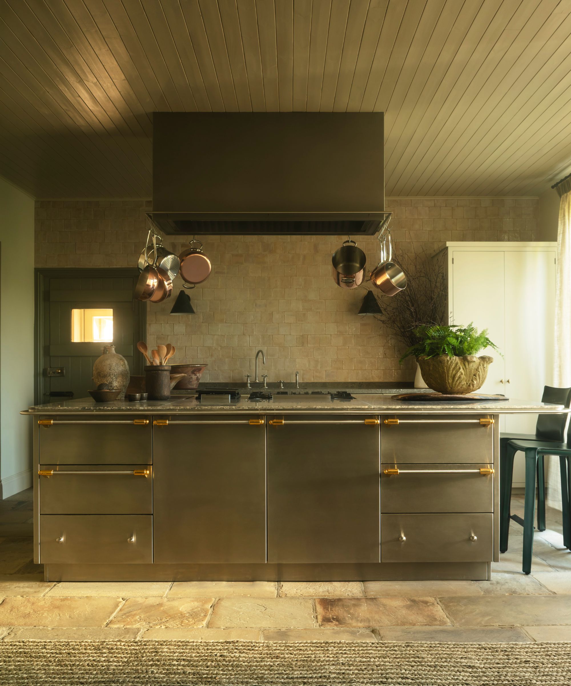 A large, industrial-style stainless steel kitchen island with brass hardware and a built-in stovetop. Copper pots hang from a dark range hood above, set against a background of textured beige stone tiles and a rustic flagstone floor.