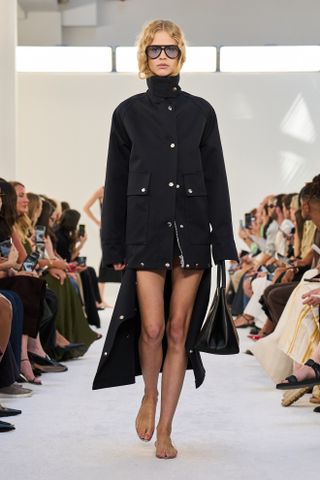 Brandon Maxwell model walking in the S/S 25 show wearing a black technical funnel-neck coat with silver buttons.