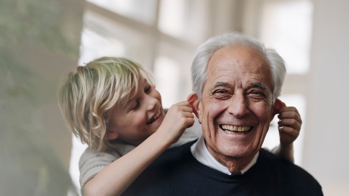 older man gives his young grandson a piggy back ride as the child laughs and gently pulls on the grandfather&#039;s ears