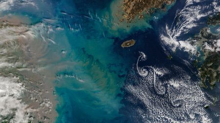 A satellite photo showing cloud swirls next to an island in the middle of the sea, as well as a massive plume of sediment extending into the water