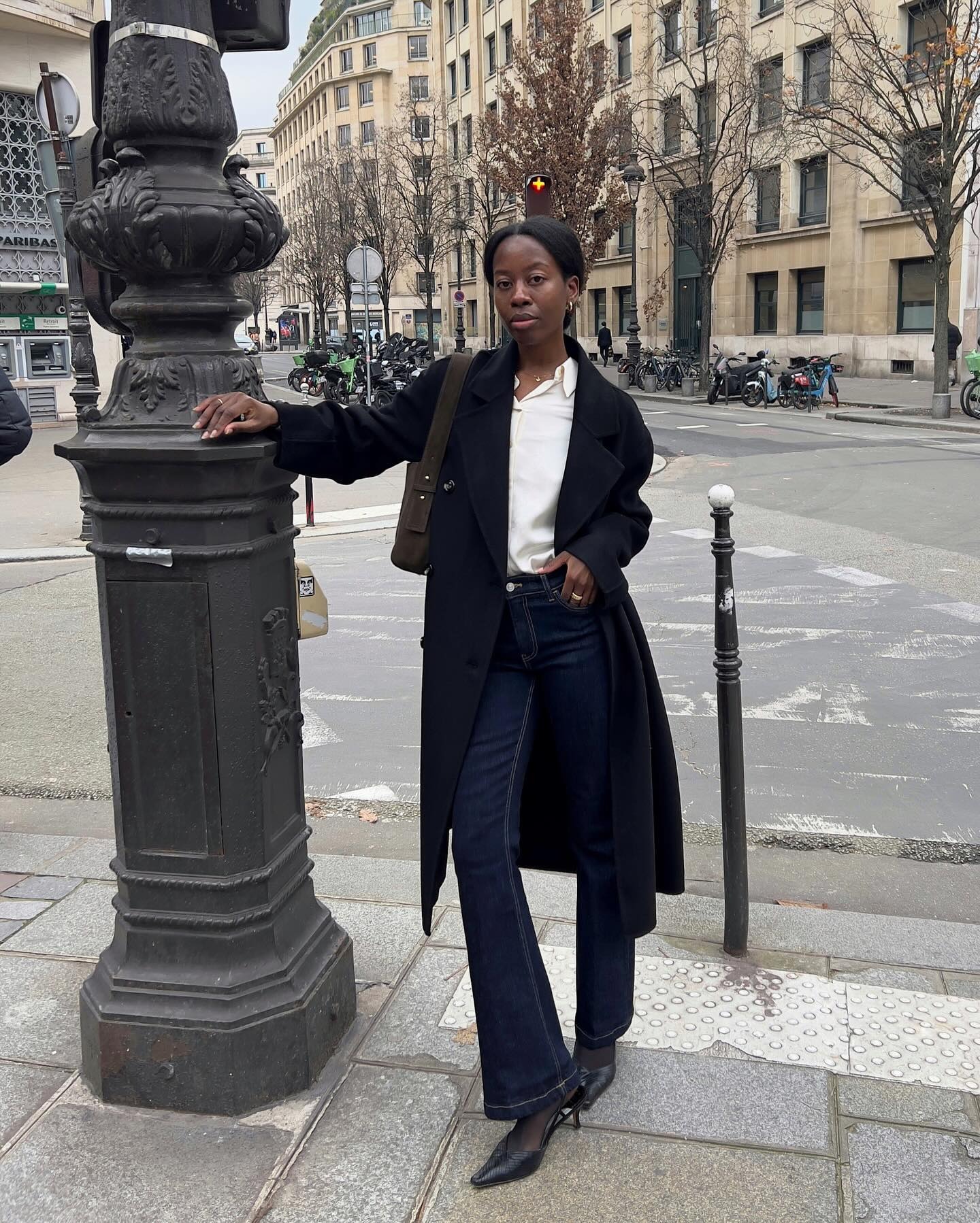 Sylvie wears a white silk button-down top, black coat, dark-wash jeans, and black slingbacks.