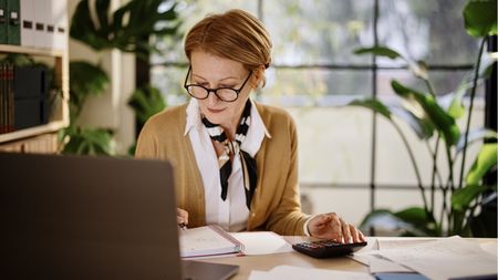 A woman looks at her laptop while using a calculator at her desk.