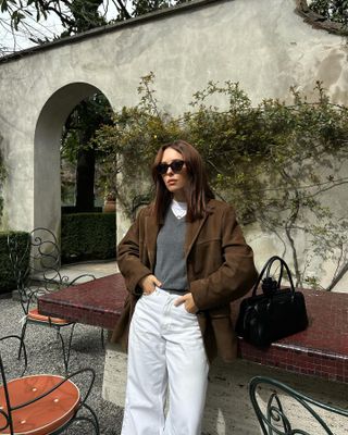 Woman wearing suede jacket, white t-shirt, gray sweater, and white jeans with black bag.