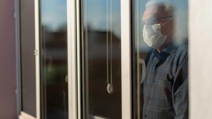 Senior man wearing COVID mask sadly stares out a window