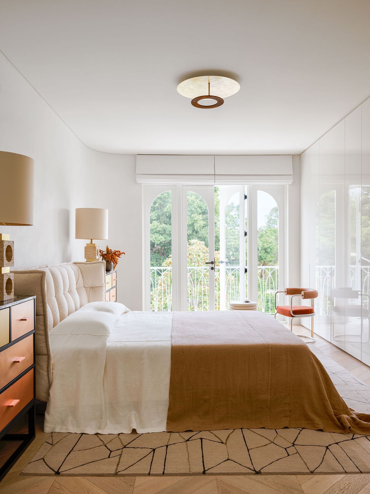 A white walled bedroom with the bed centred in the middle of the space along the left hand wall, with two chests of draws either side, and large lamps on top