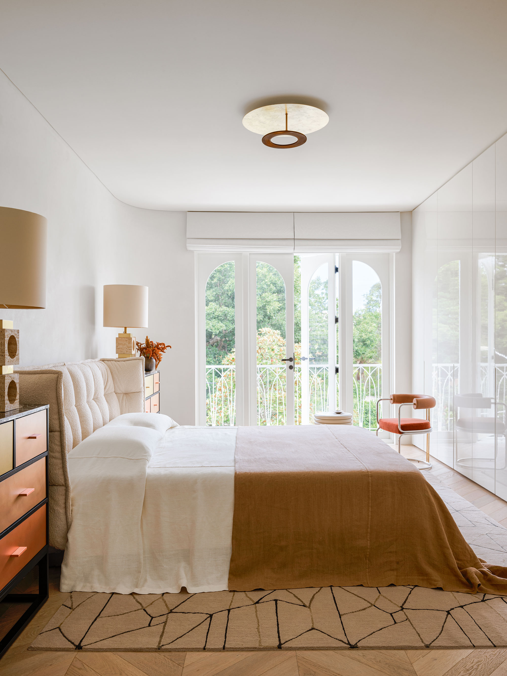 A white walled bedroom with the bed centred in the middle of the space along the left hand wall, with two chests of draws either side, and large lamps on top