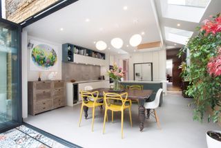 an open plan kitchen diner with bifold doors leading out to the garden - with sloping ceiling with skylights, a wooden dining table in the middle, and a kitchen island with white cabinets, and a wooden chest of drawers to the left