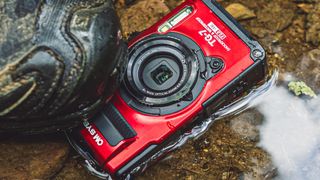 A red OM System Tough TG-7 under a boot in a puddle of water on a paved surface