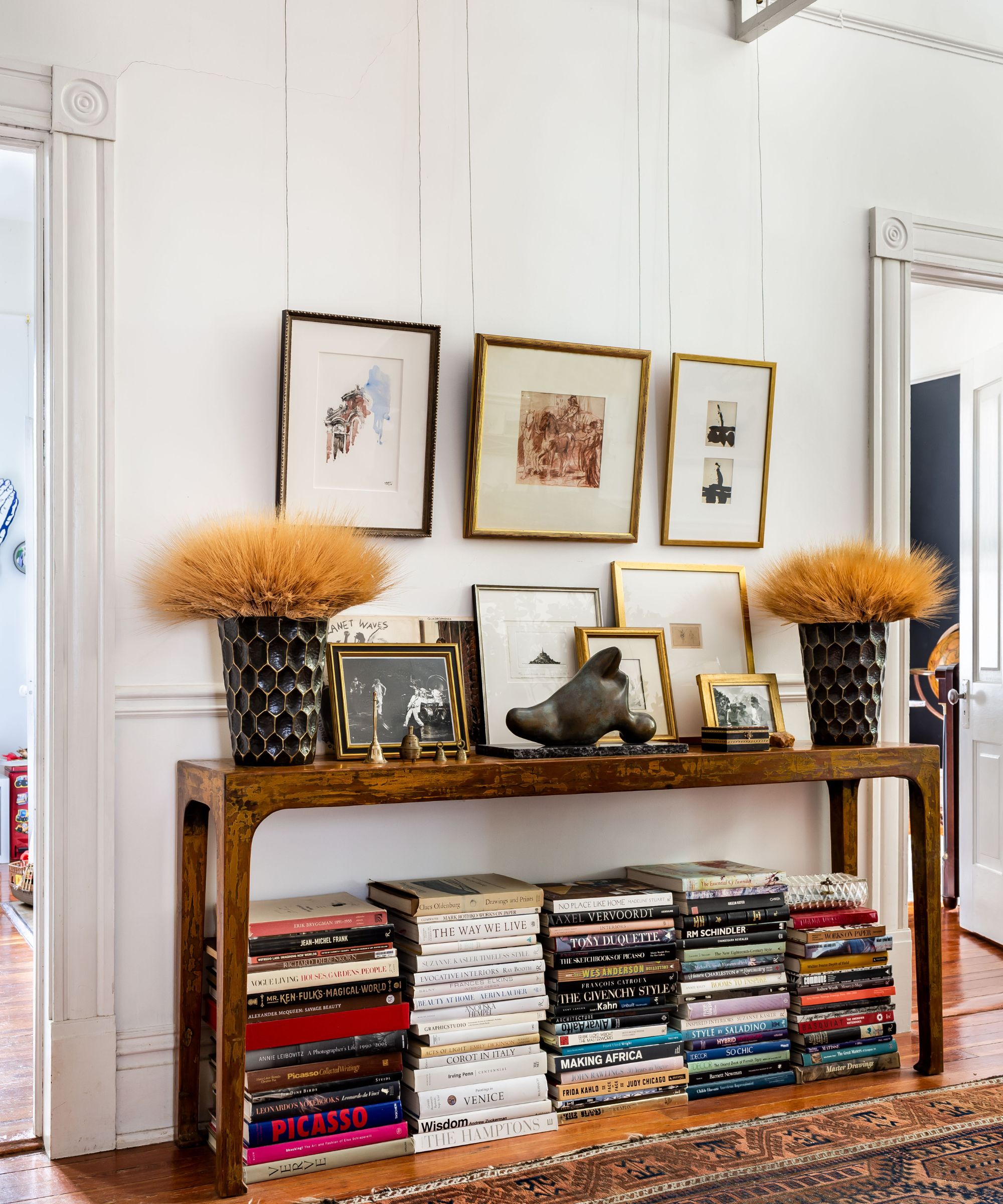 Entryway with white walls, wooden floors, a wooden console table, stacks of books underneath, and art prints hung on walls and stacked along the console table with two floral arrangements on either side