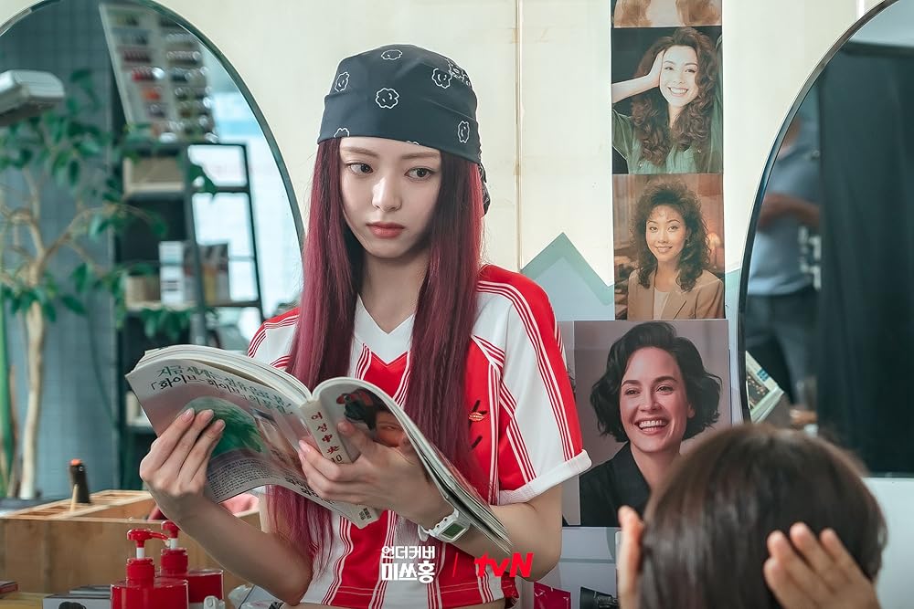 A Korean woman with red hair wearing a bandana and a red jersey (Itzy's Yuna as Hong Jang-mi) reads a magazine as she leans against the counter at hair salon, in the Netflix K-drama 'Undercover Miss Hong.'