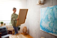 A woman looks at a canvas she prepares to hang on the wall, where another painting already hangs.