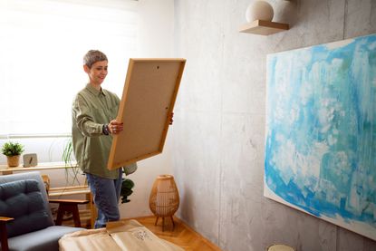 A woman looks at a canvas she prepares to hang on the wall, where another painting already hangs.
