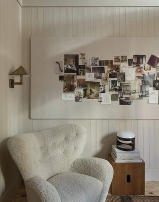 A large white boucle accent chair with a wooden side table beside it. It is sitting in the corner of an all white room with a scone light above it and a tack board with photos on it.