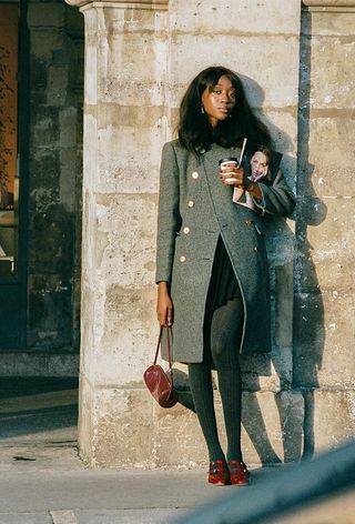 A woman wearing a long gray peacoat over a black miniskirt, gray tights, red Mary Janes, and a red handbag.