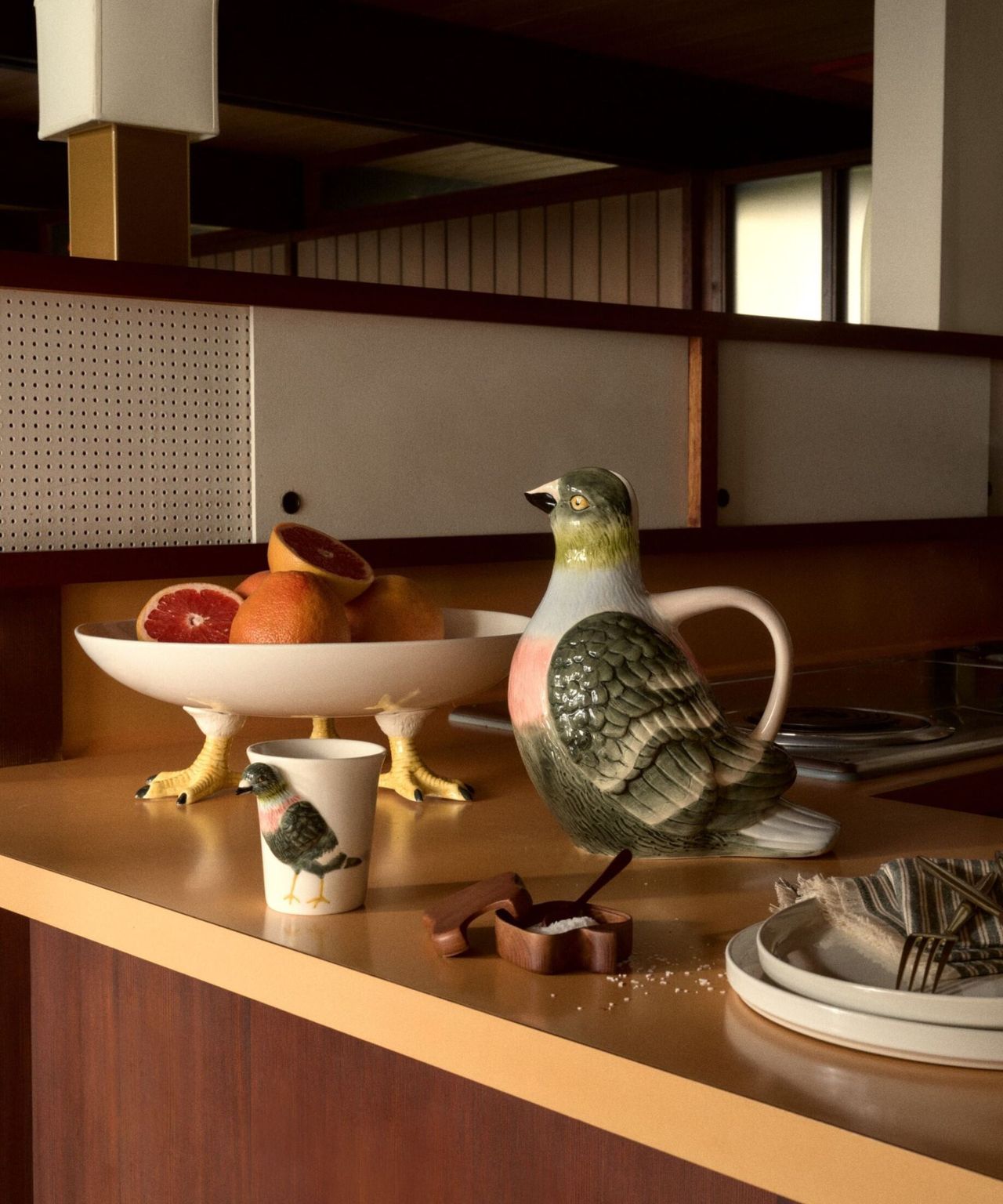 A pigeon-shaped water jug, a mug with a pigeon on it, and a serving dish with pigeon legs sitting on a mustard yellow countertop.