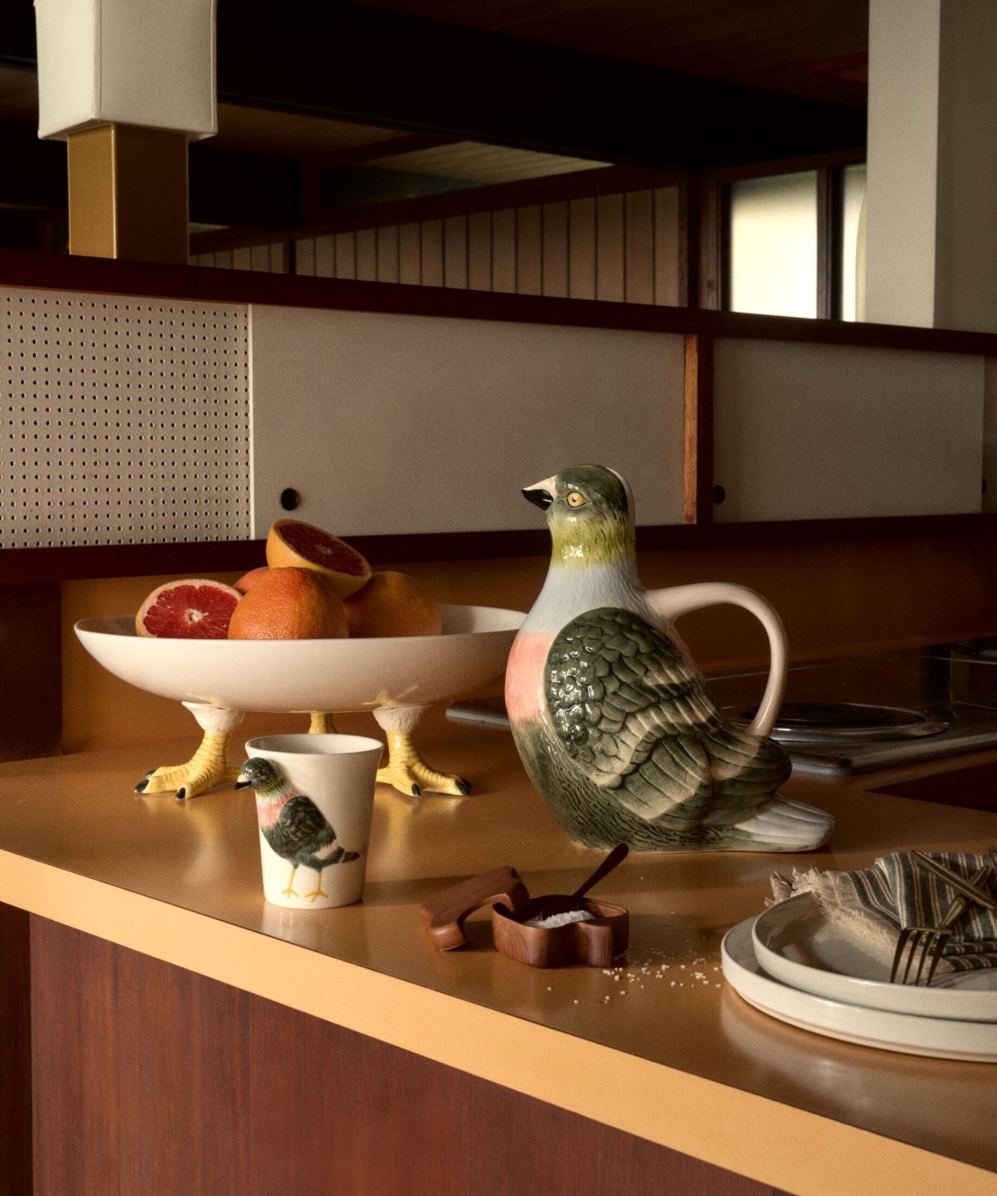 A pigeon-shaped water jug, a mug with a pigeon on it, and a serving dish with pigeon legs sitting on a mustard yellow countertop. 