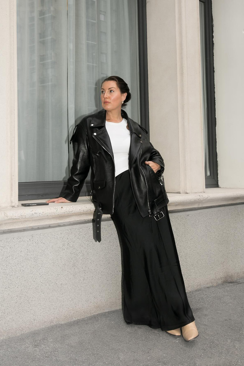 How to wear a slip skirt is shown in a woman&#039;s slip skirt outfit idea where she&#039;s leaning against a building wearing an oversized black moto jacket with a white t-shirt, black maxi satin skirt, silver earrings, and light tan moto boots
