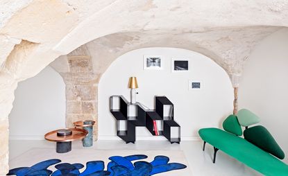 View of La Chance pieces at the Matter showroom in New York. There is a green seat with a back made up of three sections, a blue and off-white coloured rug, a black abstract shelving unit with a white and gold lamp on top and two dark coloured tables with round copper tops. The walls are off-white coloured with stone sections, there are three framed images on the wall and the ceiling is arched