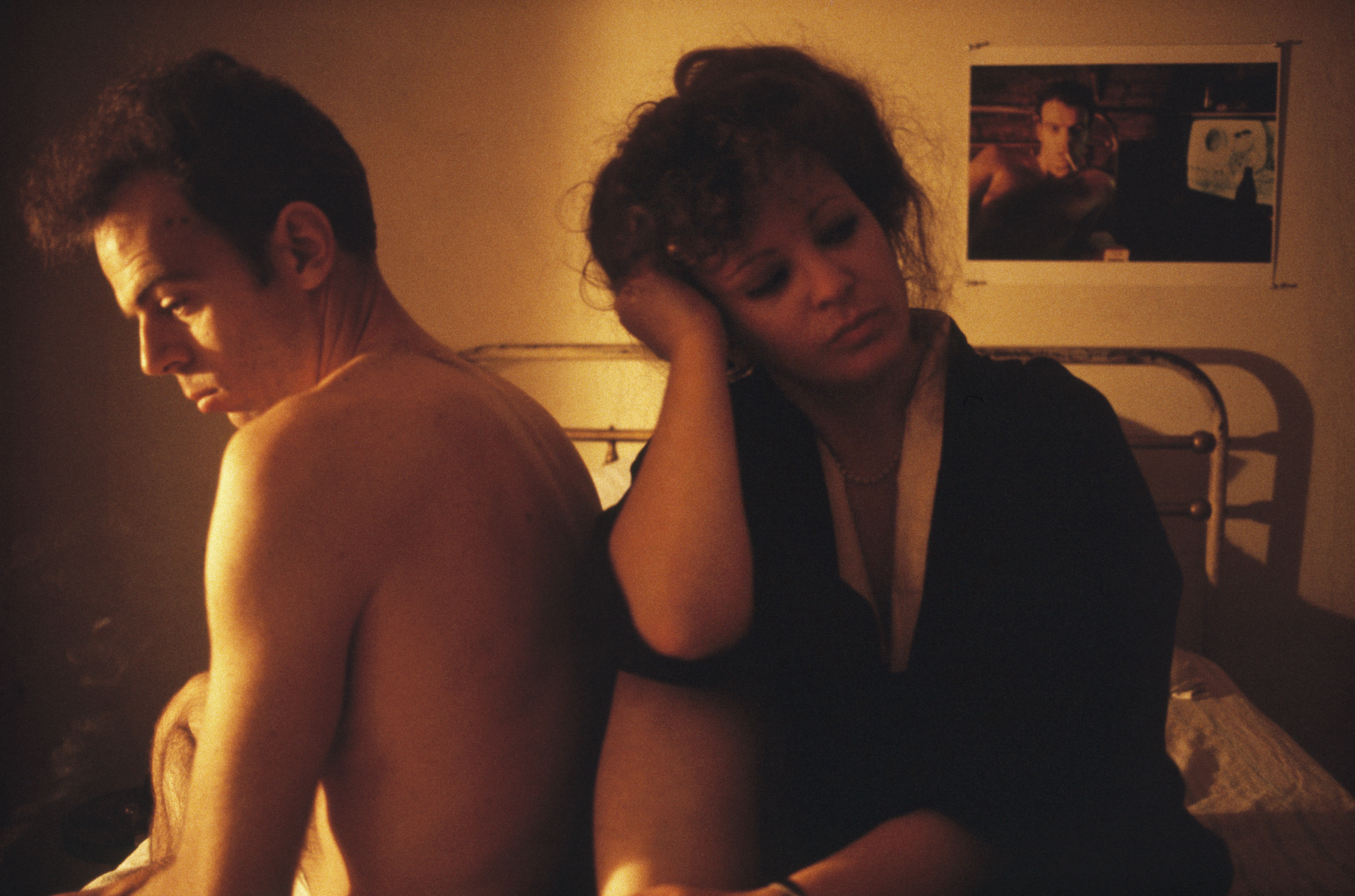 A shirtless man and a woman in a dark robe sit back to back on a bed in warm, low light, both looking downward and lost in thought.