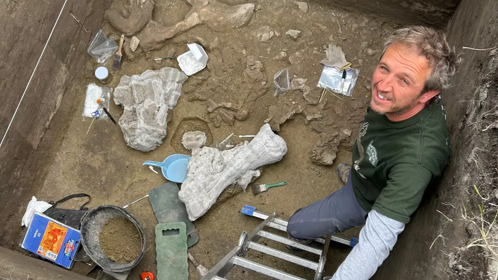 a light-skinned man with light hair and a green shirt stands at the bottom of a deep excavation looking up at the camera