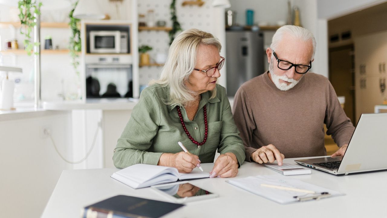 A senior couple sits at a table, working on their finances with a calculator and a computer.