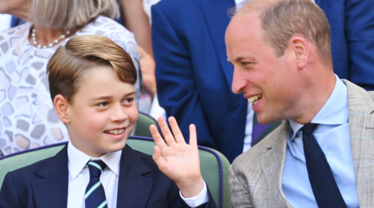 Prince George wears a navy suit and striped tie and waves while watching Wimbledon next to dad Prince William, who is wearing a gray suit