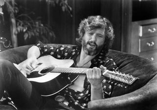 Kris Kristofferson, seated on a couch, plays an acoustic 12-string guitar