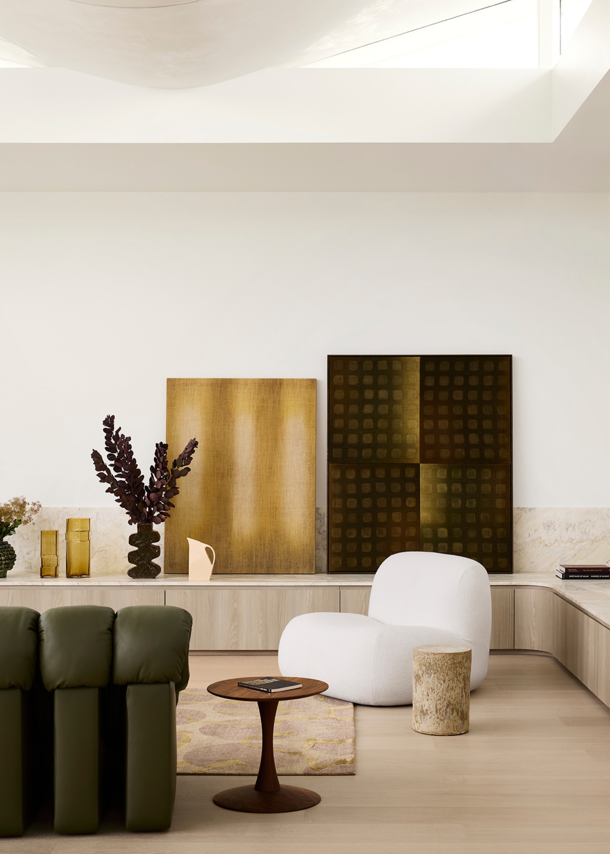 A white living room with a white accent chair, a green De Sede 'Snake' sofa, a pair of leaning paintings, a curved vase of stems, a pair of ochre vases, and a round side table