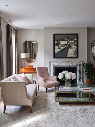 Living room with pale pink sofa and armchair beside fireplace with marbled surround, pale grey rug and mirrored coffee table