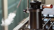 Image of a black, stovetop milk steamer on a stove with steam coming out of a silver steaming spout.