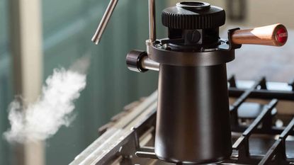 Image of a black, stovetop milk steamer on a stove with steam coming out of a silver steaming spout.