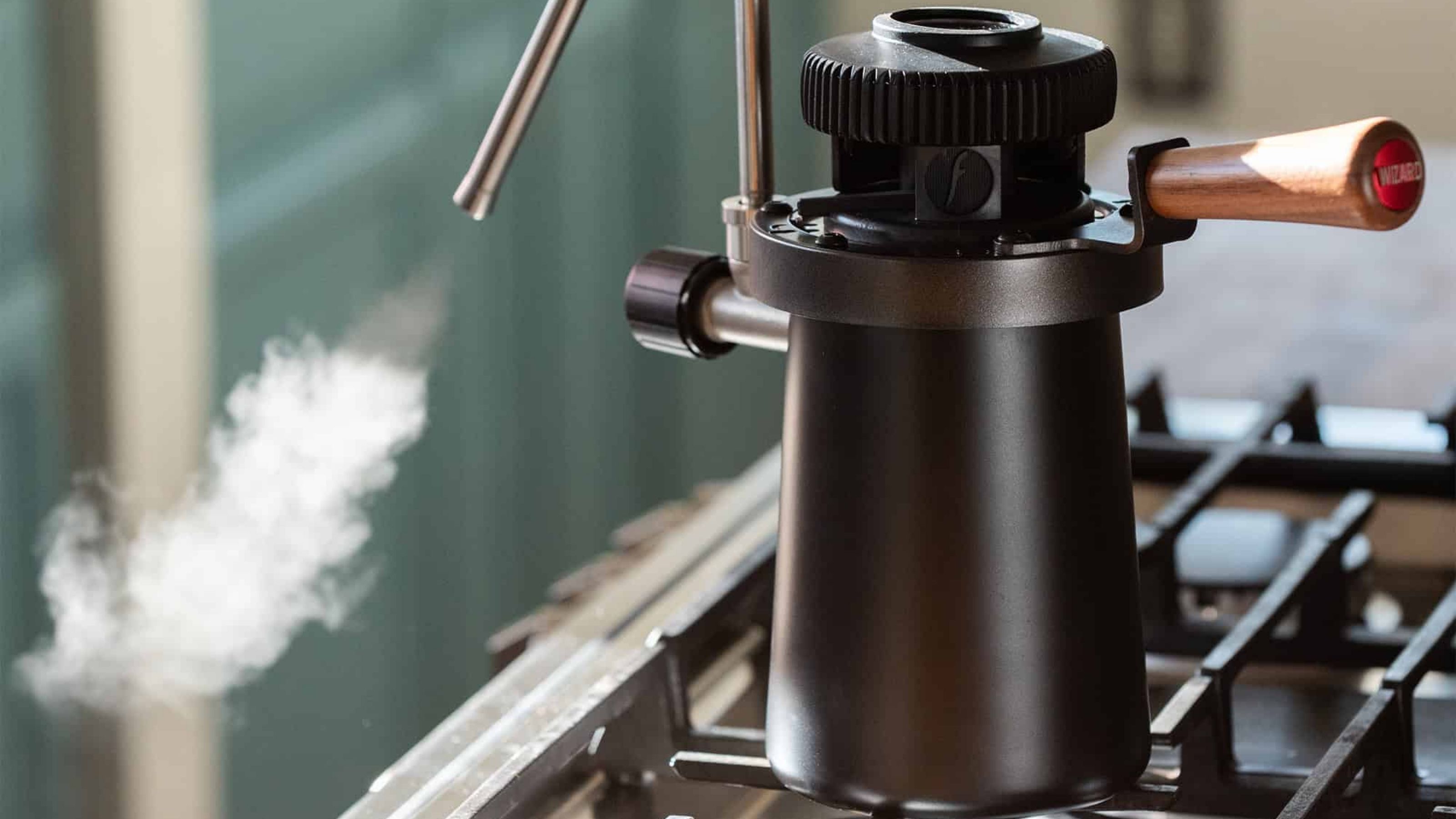 Image of a black, stovetop milk steamer on a stove with steam coming out of a silver steaming spout.