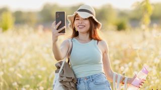 A person backlit in a sunny field wearing a hat and taking a selfie with a smartphone