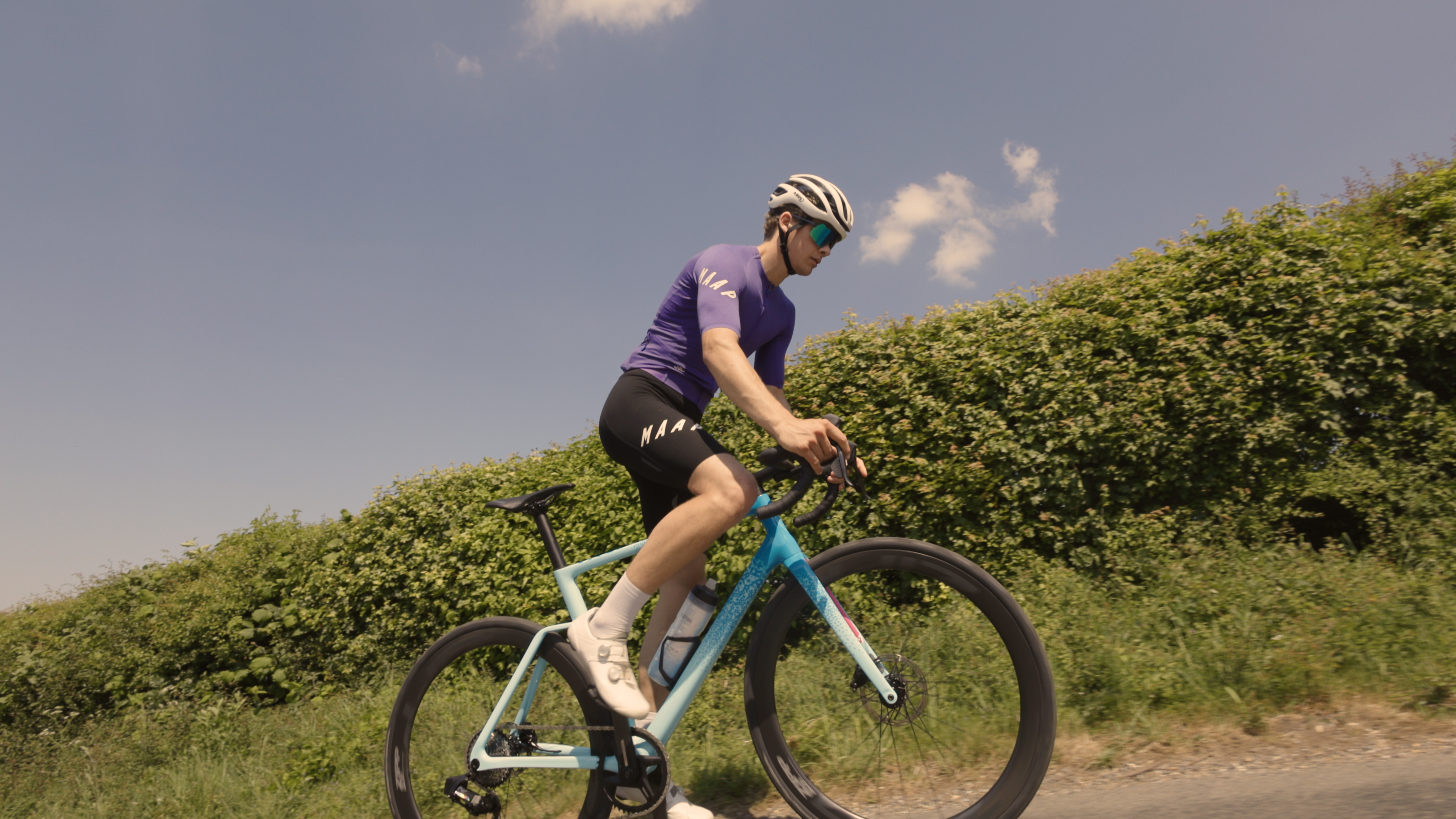 A cyclist is riding a road bike in a purple jersey and black shorts on a lane with countryside in the background