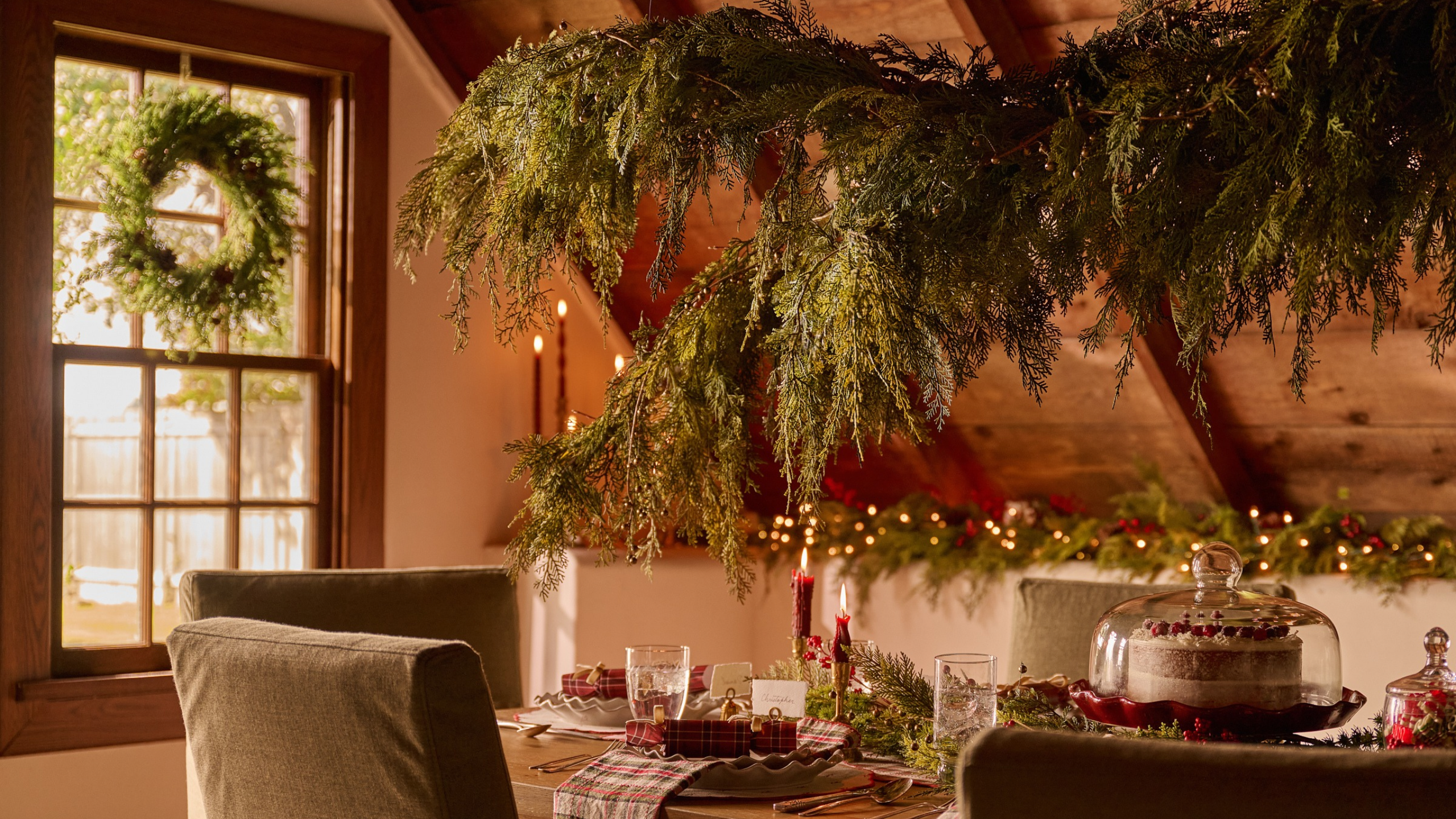 Festive Christmas dining room featuring a large evergreen garland suspended above a charmingly layered dining table set up with a cake cloche, plaid table linens, and red taper candles