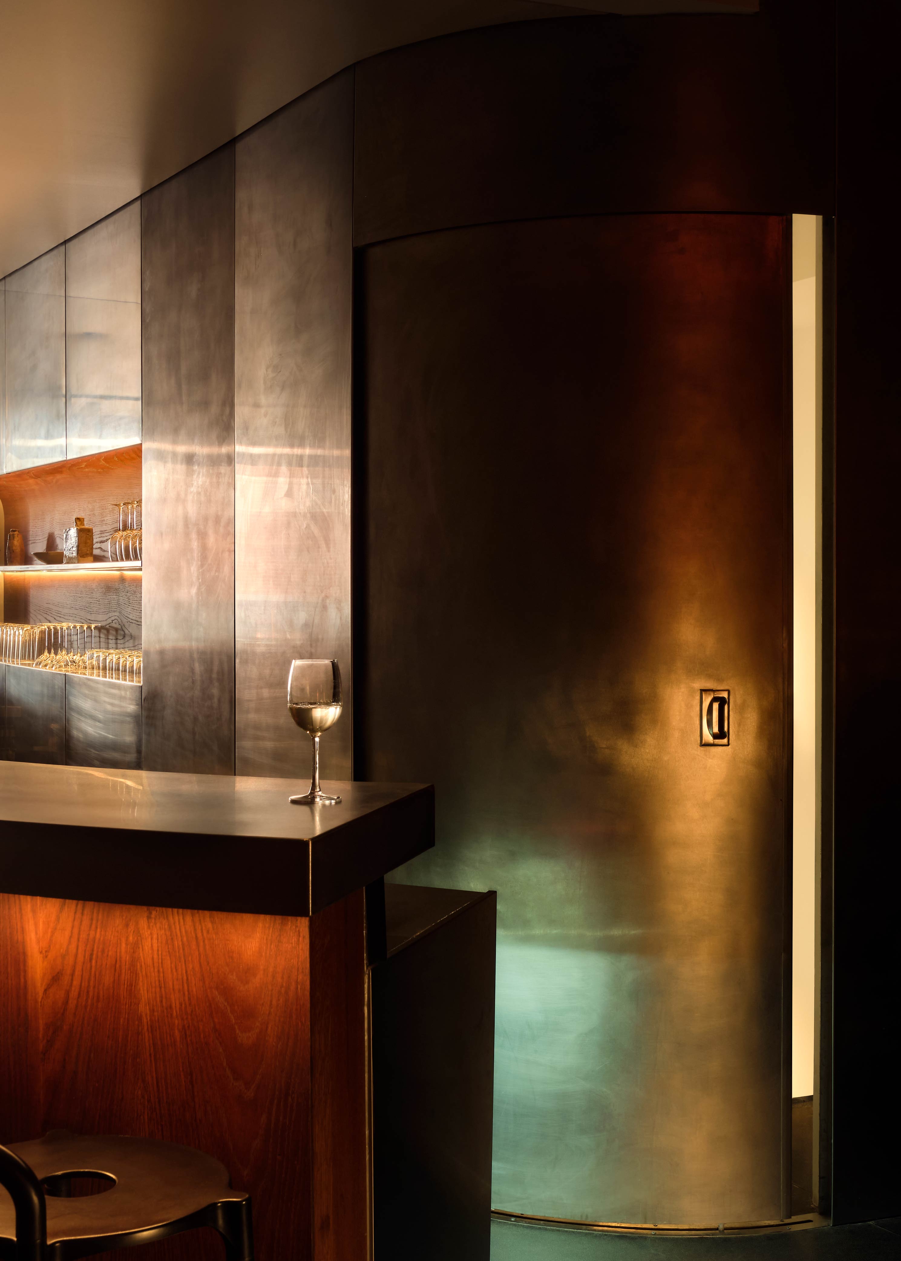 A beautifully lit bar counter, topped by a wine glass, mixes wood and chrome surfaces in a sophisticated design brought to life by gradient, soft orange, green, and yellow light.