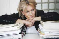 An older professional woman leans over two stacks of documents. She looks bored.