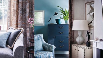 Living room with a window and floral patterned curtains, a sofa with cushions and side table. / Dark blue chest of drawers, with a small armchair in the foreground against teal painted walls. / A living room with a grey lamp and shade on a small cupboard in an alcove beside a fireplace, and long patterned curtains.