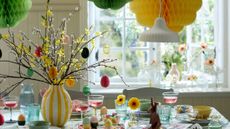 Cheerful Easter tablescape featuring a yellow floral centerpiece adorned with pastel egg ornaments, multi-colored stemware, scalloped ceramics, and a cluster of honeycomb paper decorations hanging overhead.