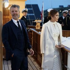 Queen Mary wearing a white polka dot dress and King Frederik in a suit standing on the royal yacht